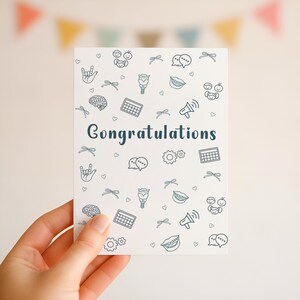 A hand holding a printable “Congratulations” card with a clean white background and SLP-themed illustrations, including AAC symbols and the ASL sign for “I love you,” ideal for celebrating new speech-language pathologists or special milestones.