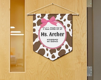 May include: A classroom banner with a cowhide pattern and a pink bow at the top. The banner reads "Y'all come on in Ms. Archer SPEECH" in a pink and brown circular design. The banner is hanging on a wooden door.