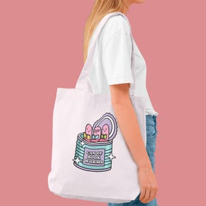 May include: White tote bag featuring a cartoon illustration of a can of worms with the text "CAN OF BOOK WORMS". The can is light blue and purple, with three smiling worms wearing glasses.