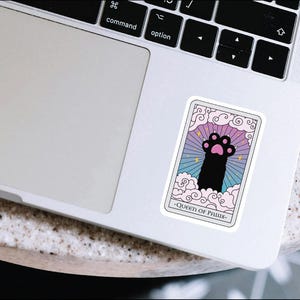 K&ouml;nnte beinhalten: Eine Tarotkarte mit einem rosa, blauen und violetten Hintergrund. Die Karte zeigt einen schwarzen Pfotenabdruck mit rosa Pfotenballen. Der Text auf der Karte lautet "Queen of Paws".