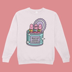 May include: A light pink sweatshirt featuring a cartoon graphic of three pink worms reading books inside a teal can. The can has the text "CAN OF BOOK WORMS" and is decorated with star shapes.