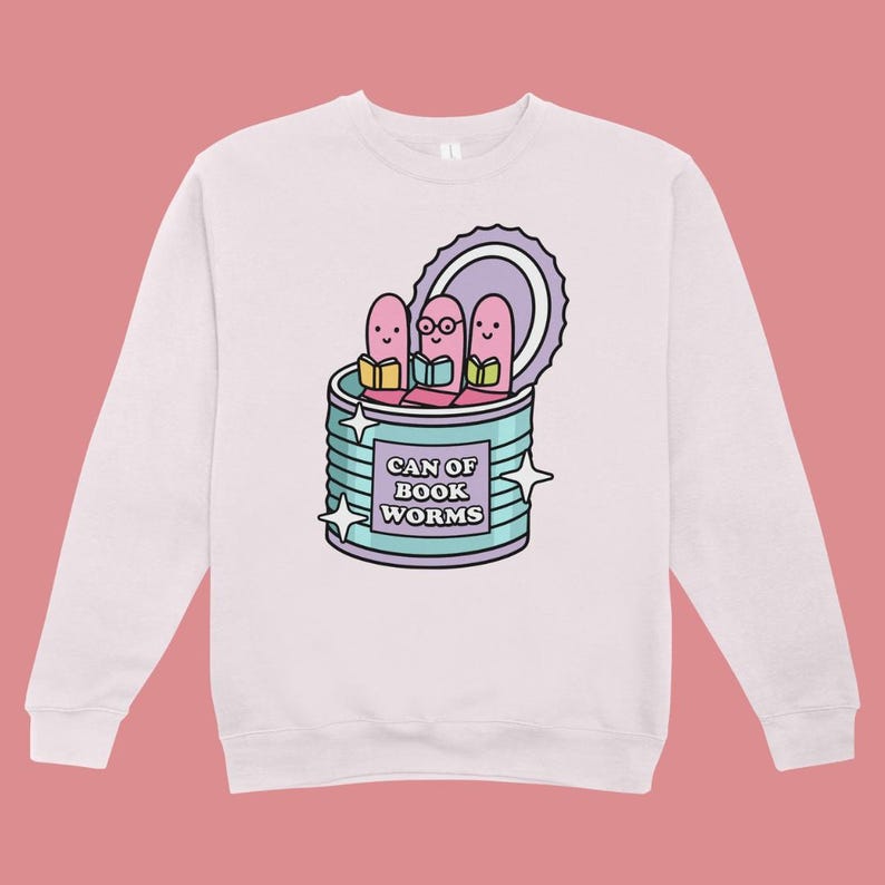 May include: A light pink sweatshirt featuring a cartoon graphic of three pink worms reading books inside a teal can. The can has the text "CAN OF BOOK WORMS" and is decorated with star shapes.