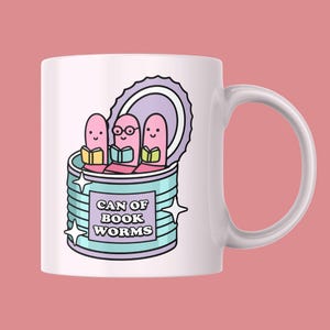 May include: White ceramic mug featuring a whimsical illustration of three pink worm characters reading books inside a can. The can is light blue with the text "CAN OF BOOK WORMS" and star accents. The background is a solid pink.