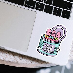 May include: A sticker featuring a whimsical design of three pink bookworms reading inside a teal can. The can is labeled "CAN OF BOOK WORMS" in purple text, with star accents. The sticker is placed on a laptop.