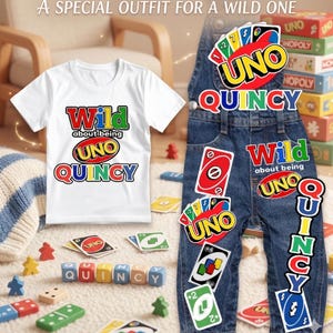 Puede incluir: Una camiseta blanca y un peto vaquero con gráficos coloridos con temática de UNO y el nombre "Quincy". La camiseta dice "Wild about being UNO Quincy". El peto está decorado con cartas y texto de UNO. La imagen también incluye piezas del juego UNO.