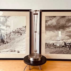 May include: A silver table lamp with a black base and three legs. The lamp has a simple design with a round shade and a vertical stem.