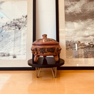 May include: A terracotta-colored, lidded container with carved details, resting on a small, black, round stand with gold-colored hairpin legs. The container features sculpted faces and patterns. Two framed black and white landscape prints are in the background.