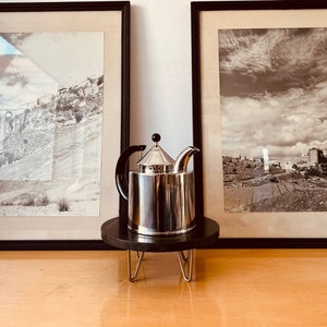 May include: A silver teapot with a black handle and lid sits on a black wooden stand with gold metal legs. The teapot is in front of two black framed pictures with black and white photos.