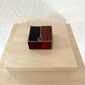 May include: A small, stained glass box with a geometric design. The top is divided into black and reddish-brown sections, with a small, round, reddish-brown gem in the center. The bottom section is red. The box is sitting on a light-colored wooden surface.
