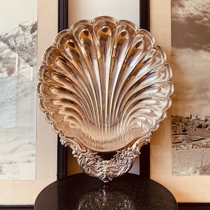 May include: A silver shell-shaped serving dish with ornate details. The dish is displayed on a black pedestal.