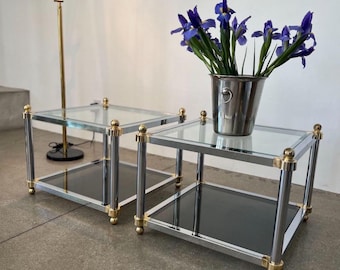 May include: Two chrome and brass end tables with glass shelves. The tables are square and have a mirrored bottom shelf. A bouquet of purple irises is in a silver bucket on top of one of the tables.