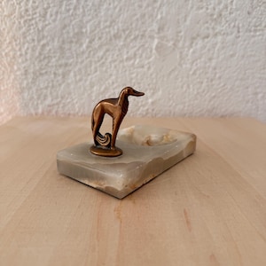 May include: A bronze-colored dog figurine atop a rectangular, light-colored stone ashtray. The dog is slender with a curved tail. The ashtray has a small, circular indentation. The piece is set on a light-colored wooden surface.