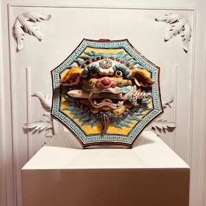 May include: A colorful ceramic wall plaque depicting a mythical lion with a sword in its mouth. The plaque is octagonal and features a yellow background with blue and green accents.