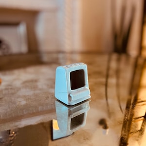 May include: A small, white, plastic model of a vintage computer. The model has a black screen and a few buttons on the front.