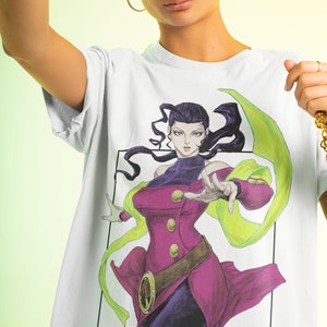 May include: White t-shirt with a colourful illustration of a character in a magenta and purple outfit with a green scarf. The artwork is framed in a black border, with Japanese text at the bottom. The character has dark hair and is in a dynamic pose.