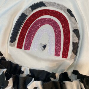 May include: A white long-sleeved shirt with a rainbow design. The rainbow features red, pink, and white glitter, with cow print and black leather accents. The bottom of the shirt has a black and white cow print ruffle.
