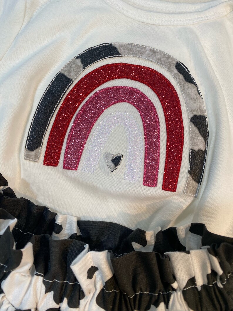 May include: A white long-sleeved shirt with a rainbow design. The rainbow features red, pink, and white glitter, with cow print and black leather accents. The bottom of the shirt has a black and white cow print ruffle.