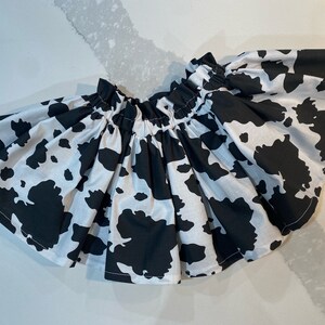May include: A white and black cow print skirt. The skirt is gathered at the waist and flares out. The fabric has large, irregular black spots on a white background. The skirt is likely for a child.