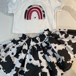 May include: A white baby bodysuit with a rainbow design in gray, red, and pink glitter, paired with a black and white cow print skirt. The skirt is gathered at the waist.