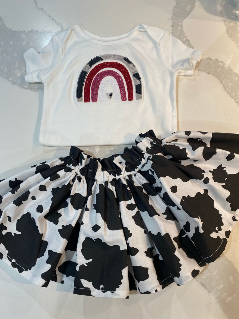 May include: A white baby bodysuit with a rainbow design in gray, red, and pink glitter, paired with a black and white cow print skirt. The skirt is gathered at the waist.