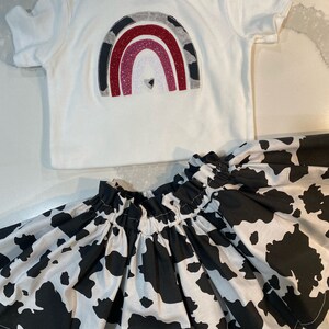 May include: A white baby bodysuit with a rainbow design in shades of gray, red, and glitter. Paired with a black and white cow print skirt. The outfit is designed for infants and toddlers.