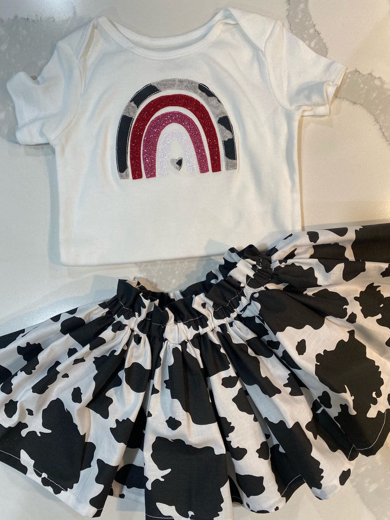 May include: A white baby bodysuit with a rainbow design in shades of gray, red, and glitter. Paired with a black and white cow print skirt. The outfit is designed for infants and toddlers.