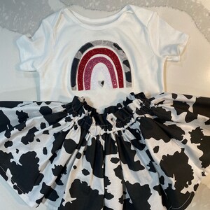 May include: A white baby bodysuit with a rainbow design and a black and white cow print skirt. The rainbow has gray, red, and glittery pink arches. A small heart is in the center of the rainbow.