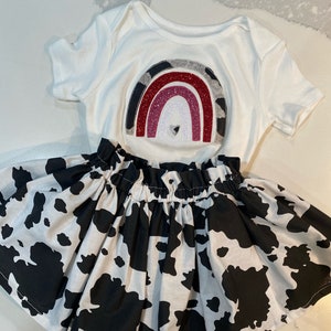 May include: A white baby bodysuit with a rainbow design and a black and white cow print skirt. The rainbow has a heart in the center and is made of glitter and other materials. The skirt is gathered at the waist.