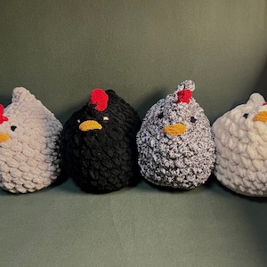 May include: Four crocheted chicken plushies in different colors: white, black, gray, and white with black speckles. Each plush has a red comb and a yellow beak.