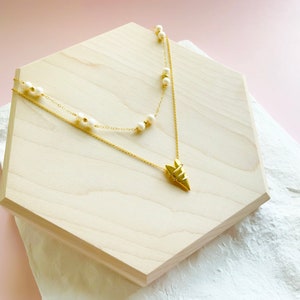 May include: A gold necklace with a geometric pendant and a strand of white pearls. The necklace is displayed on a wooden hexagon.
