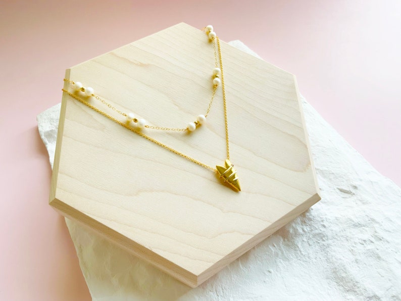 May include: A gold necklace with a geometric pendant and a strand of white pearls. The necklace is displayed on a wooden hexagon.