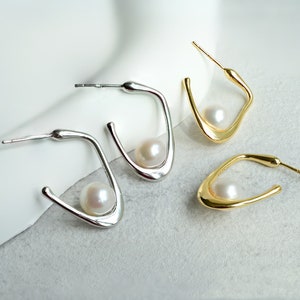 May include: A pair of silver and gold hoop earrings with a pearl accent. The earrings are in the shape of a curved line.