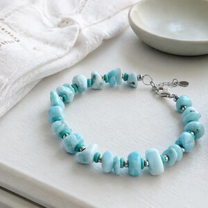 Puede incluir: Una pulsera que presenta piedras de Larimar azul claro, irregulares y pulidas, intercaladas con peque&ntilde;as cuentas met&aacute;licas cil&iacute;ndricas de tono plateado y diminutas cuentas espaciadoras de color turquesa, con un cierre de mosquet&oacute;n plateado y una peque&ntilde;a etiqueta circular.
