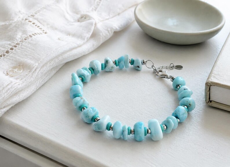 Puede incluir: Una pulsera que presenta piedras de Larimar azul claro, irregulares y pulidas, intercaladas con peque&ntilde;as cuentas met&aacute;licas cil&iacute;ndricas de tono plateado y diminutas cuentas espaciadoras de color turquesa, con un cierre de mosquet&oacute;n plateado y una peque&ntilde;a etiqueta circular.