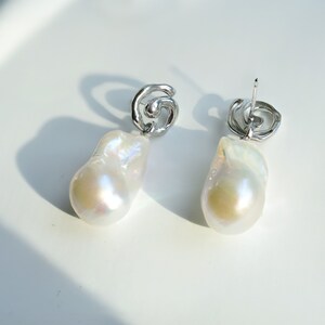 May include: A pair of drop earrings featuring large, irregular white baroque pearls suspended from silver-toned metal studs. The studs have a swirling, organic shape, and the pearls exhibit a lustrous, iridescent surface with subtle pink and blue tones.