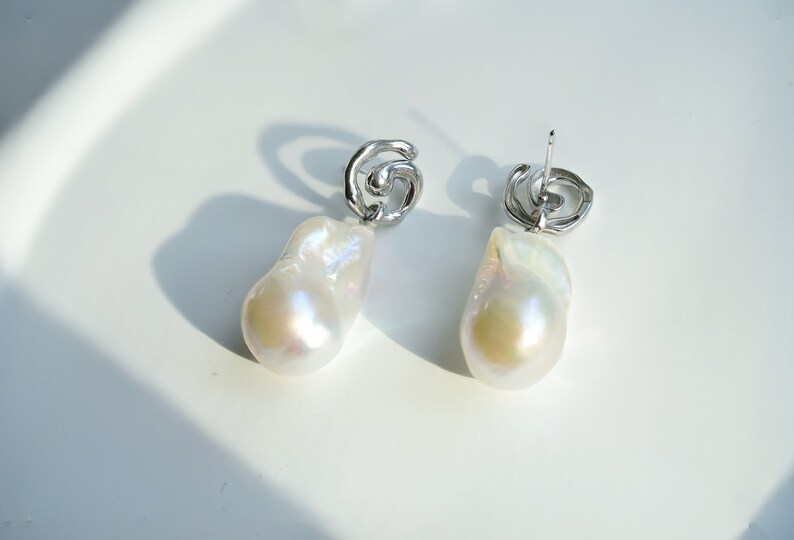 May include: A pair of drop earrings featuring large, irregular white baroque pearls suspended from silver-toned metal studs. The studs have a swirling, organic shape, and the pearls exhibit a lustrous, iridescent surface with subtle pink and blue tones.