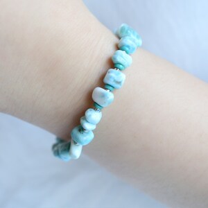 Puede incluir: Una pulsera de cuentas que presenta piedras pulidas de color azul claro y blanco veteado, intercaladas con peque&ntilde;as cuentas redondas de color turquesa vibrante y finos detalles met&aacute;licos en tono plateado.