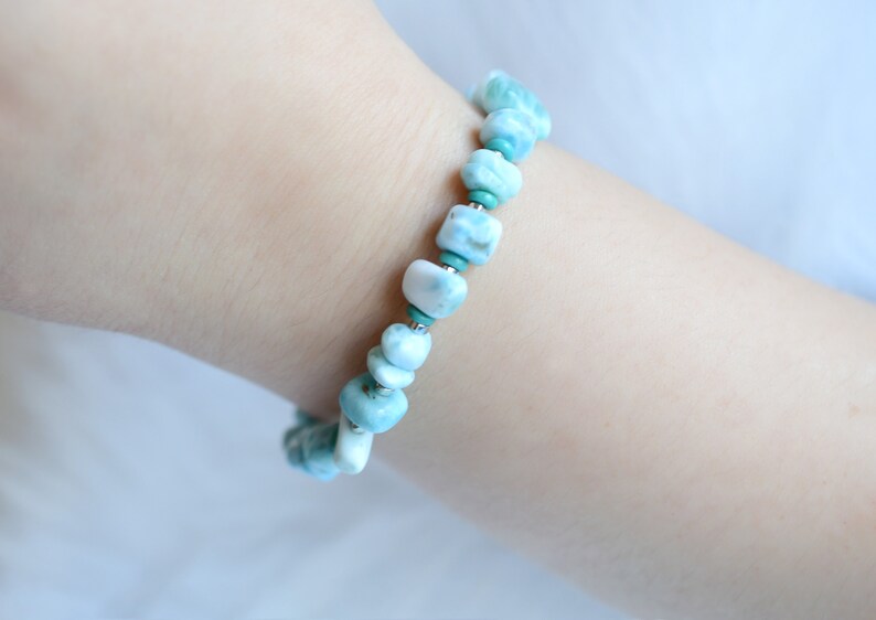 Puede incluir: Una pulsera de cuentas que presenta piedras pulidas de color azul claro y blanco veteado, intercaladas con peque&ntilde;as cuentas redondas de color turquesa vibrante y finos detalles met&aacute;licos en tono plateado.