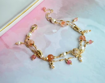 May include: A delicate gold bracelet with two strands of chain and a variety of beads, including pink, white, and orange. The bracelet features a small, ornate charm.