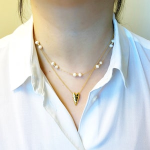 May include: A gold necklace with a geometric pendant and a pearl necklace layered on top. The necklaces are worn by a person with a white shirt.