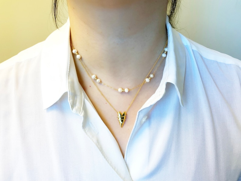 May include: A gold necklace with a geometric pendant and a pearl necklace layered on top. The necklaces are worn by a person with a white shirt.