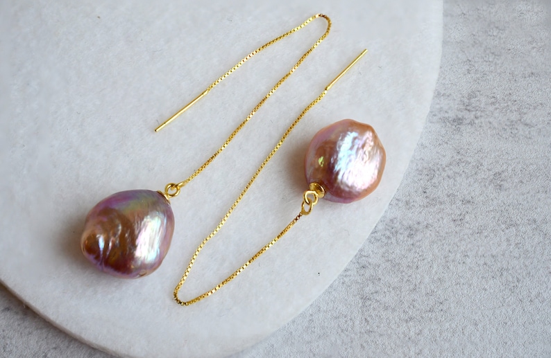 May include: A pair of gold chain earrings with iridescent pink baroque pearls. The pearls are teardrop shaped and dangle from the chains.
