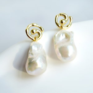 May include: A pair of gold-tone earrings featuring a swirling, open-work metal design at the top, from which a large, irregular, white baroque pearl hangs. The pearls have a lustrous, iridescent finish and a smooth, organic, non-uniform shape.