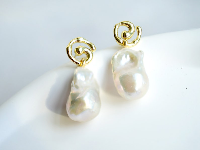 May include: A pair of gold-tone earrings featuring a swirling, open-work metal design at the top, from which a large, irregular, white baroque pearl hangs. The pearls have a lustrous, iridescent finish and a smooth, organic, non-uniform shape.