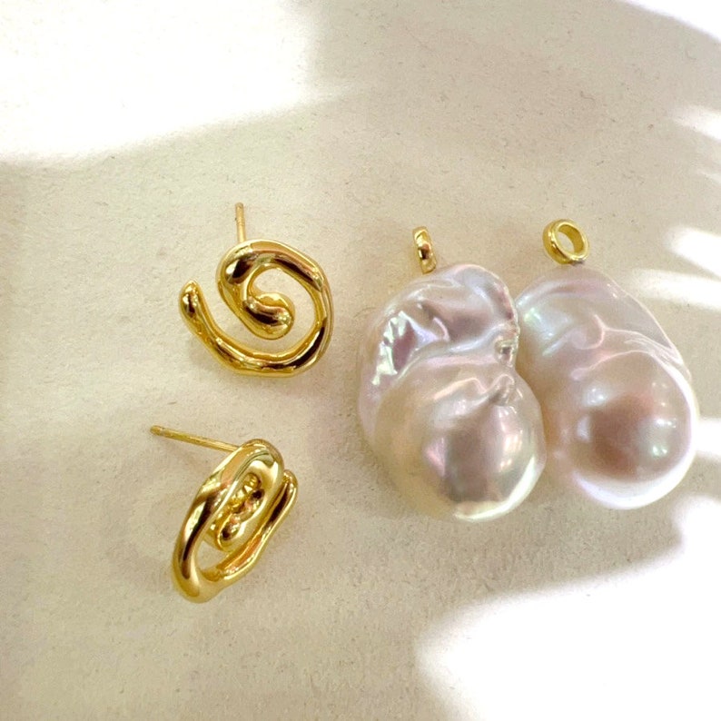 May include: Two gold-toned metal stud earrings with an open, swirling, organic wire design alongside two large, irregular, iridescent white baroque pearls, each featuring a small gold-toned metal loop at the top for attachment as pendants.