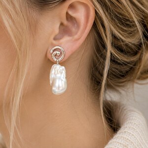 May include: A silver-toned earring featuring a swirling, textured metal stud top connected to a large, irregular white baroque pearl drop. The earring is designed with a polished finish that catches the light, highlighting the organic shape of the pearl.