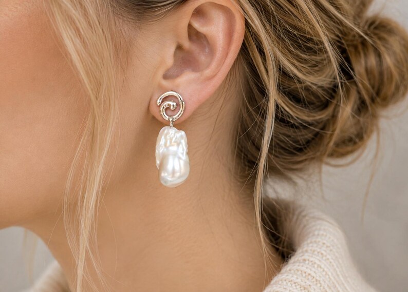 May include: A silver-toned earring featuring a swirling, textured metal stud top connected to a large, irregular white baroque pearl drop. The earring is designed with a polished finish that catches the light, highlighting the organic shape of the pearl.