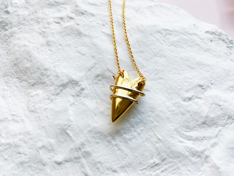 May include: A gold necklace with a heart-shaped pendant. The pendant is made of two pieces of metal, one gold and one silver, that are intertwined. The necklace is on a white background.