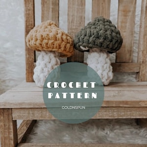 May include: Two crocheted mushroom amigurumi toys in mustard yellow and forest green with white stems. The mushrooms are sitting on a wooden bench with a white fuzzy rug underneath. The text "CROCHET PATTERN GOLDNSPUN" is on a teal circle in the foreground.