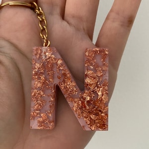 May include: A pink resin letter N keyring with gold flecks. The keyring is attached to a gold chain.